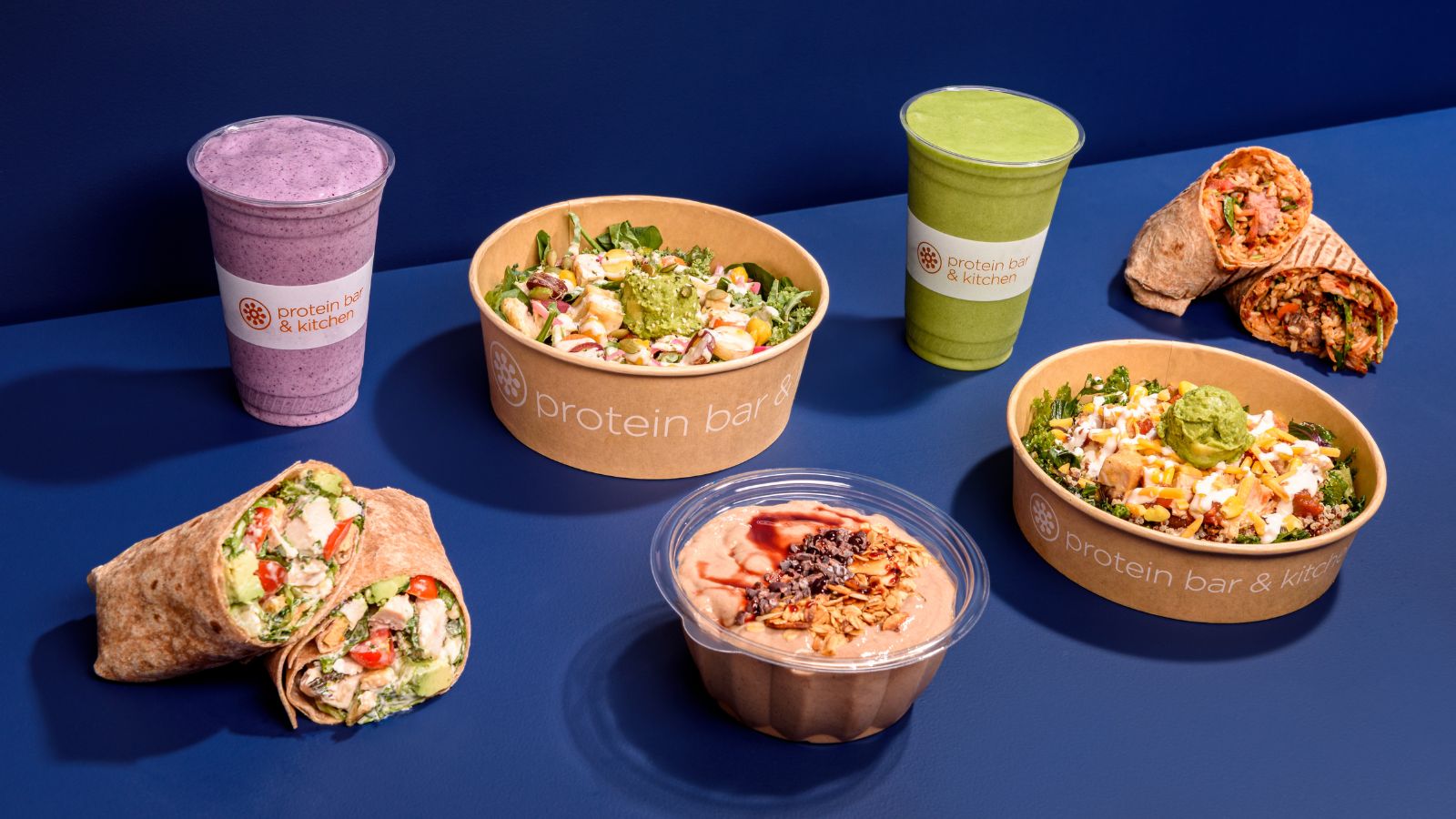 Protein Bar & Kitchen bowls, wraps, smoothies and acai bowl arranged on a navy table